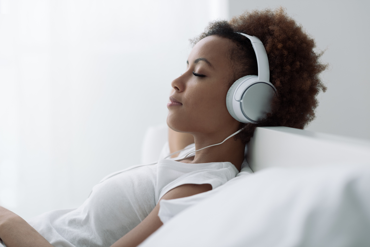 woman laying down in a white shirt on a white bed listening to music through white headphones.