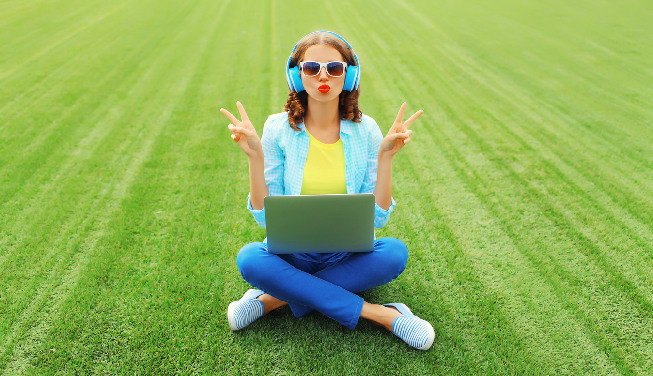 Woman sitting in a field with her laptop. SHe's wearing headphones and holding up two peace signs with her hands.