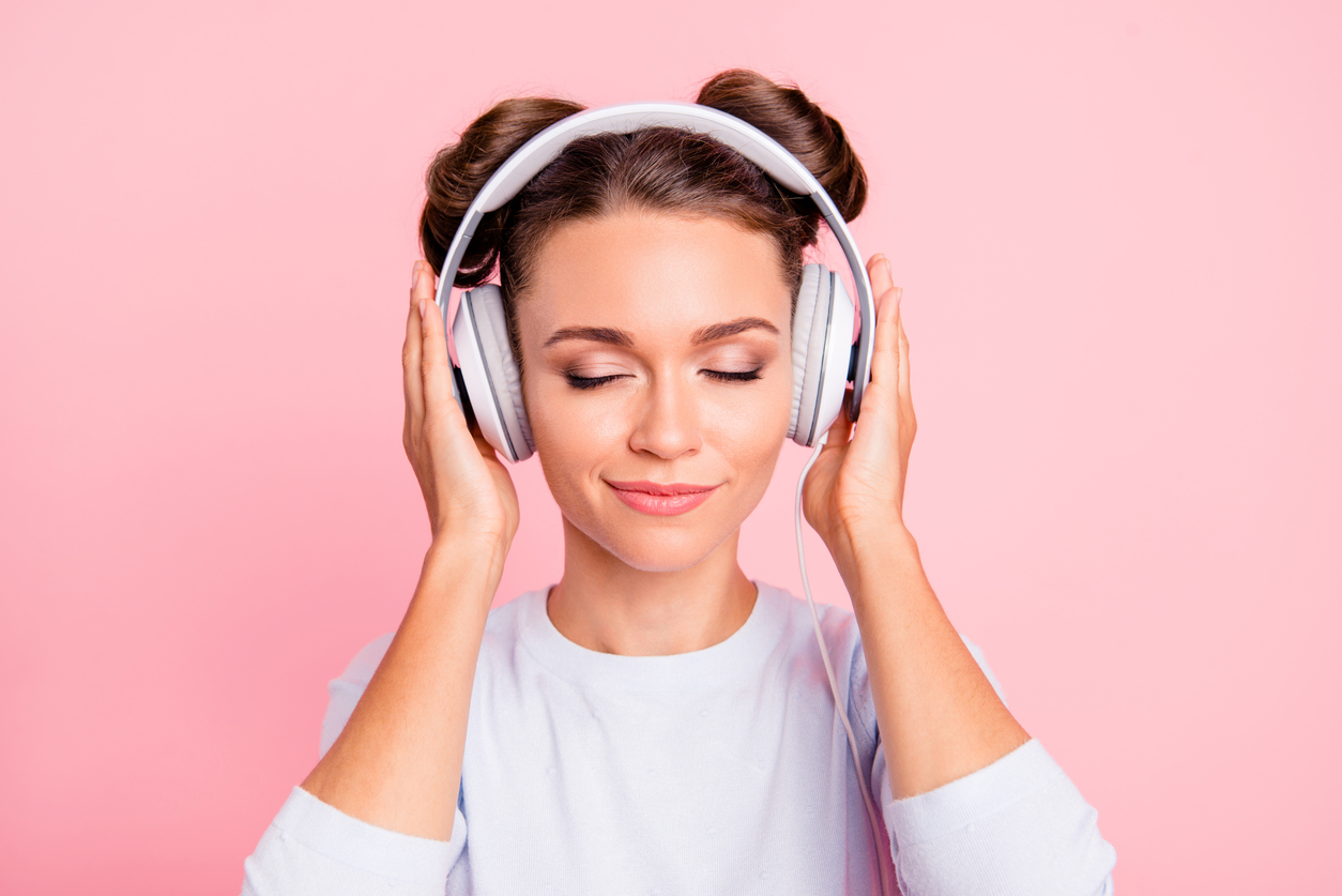 Woman with eyes closed wearing white headphones on a pink background.